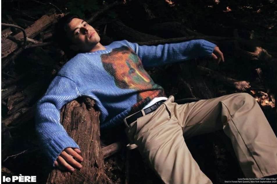 A young man rests in a forest, showcasing fashion with a blue patterned sweater and khaki pants, illuminated by a soft, natural light.