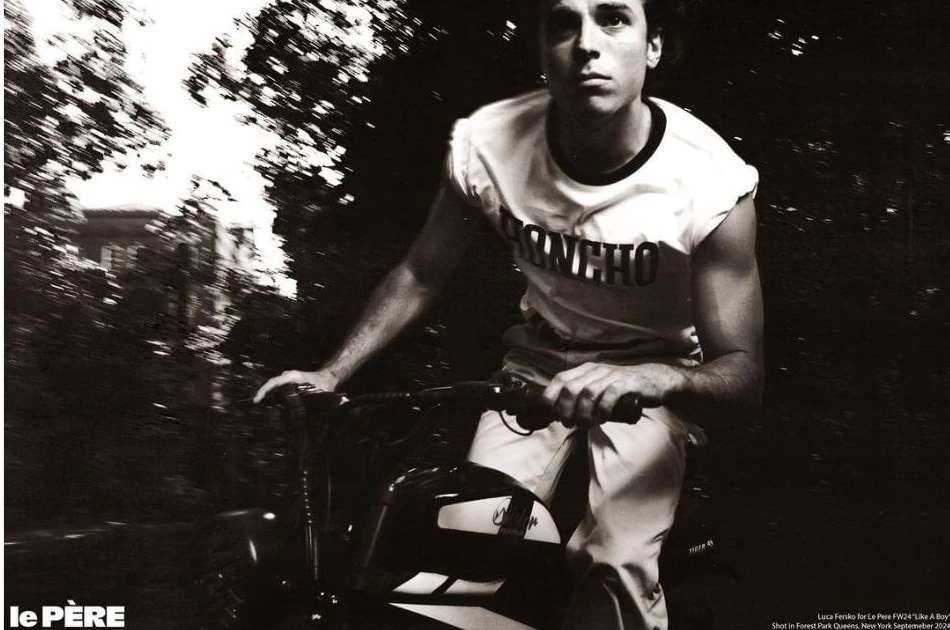 Monochromatic shot of a young adult male riding a bike outdoors on a path surrounded by trees, wearing casual athletic clothing.