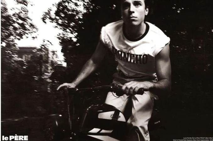 Monochromatic shot of a young adult male riding a bike outdoors on a path surrounded by trees, wearing casual athletic clothing.
