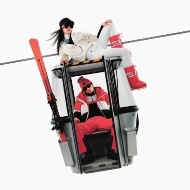 Two people in ski outfits on a cable car, with one person seated inside and the other sitting atop the car roof.