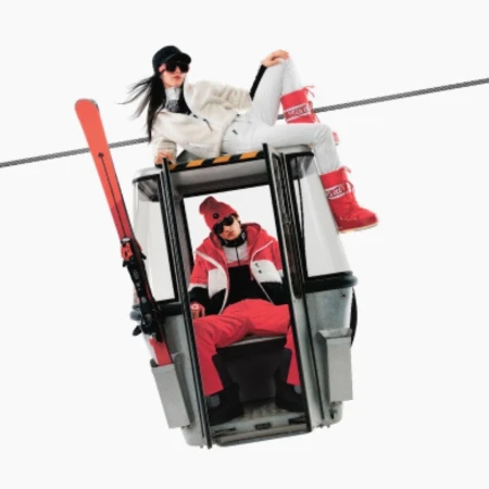 Two people in ski outfits on a cable car, with one person seated inside and the other sitting atop the car roof.