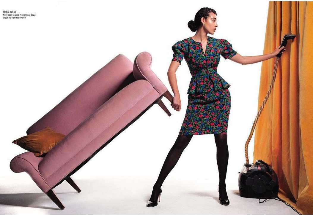 A stylish woman in a floral dress holds a vintage vacuum cleaner and poses next to a modern pink sofa in a bright photo studio.