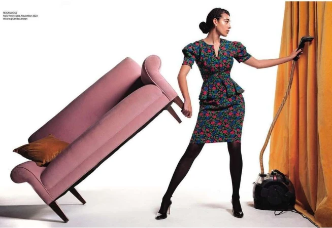 A stylish woman in a floral dress holds a vintage vacuum cleaner and poses next to a modern pink sofa in a bright photo studio.