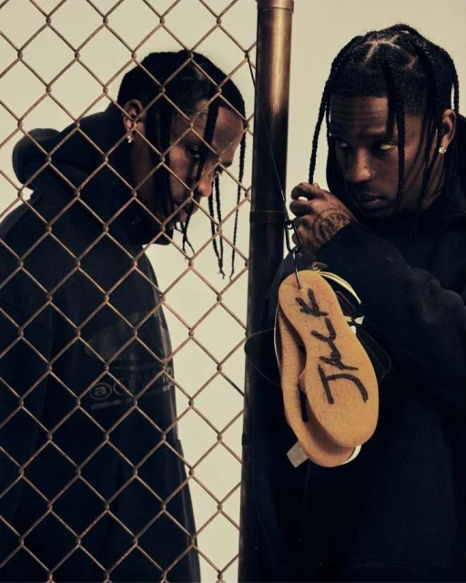 Two adult males, one holding shoe insoles with writing, stand near a chain-link fence in a studio, showcasing streetwear fashion.