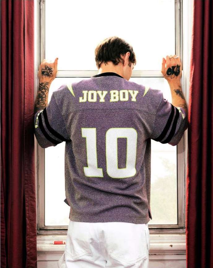 Back view of a man in a vintage football jersey looking out a window, hand tattoos visible, bright indoor light with red curtains.