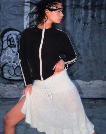 Model with dark hair blowing in the wind wearing a black zip jacket and white skirt against a concrete wall