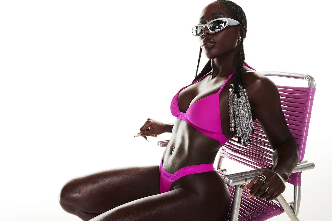 An African American adult female model poses while sitting on a pink lawn chair in a bright pink bikini and futuristic sunglasses.