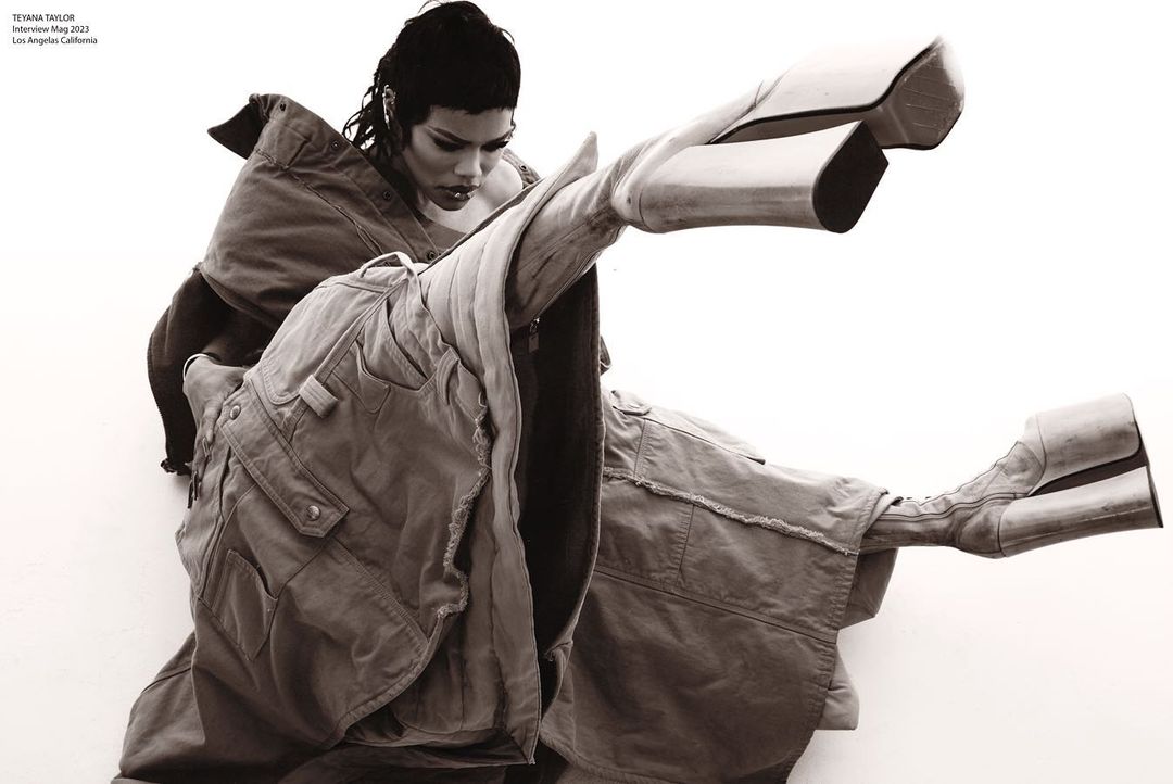 Fashion editorial shot featuring a model in an avant-garde pose, showcasing a large cargo-style jacket and oversized boots against a clean white backdrop.