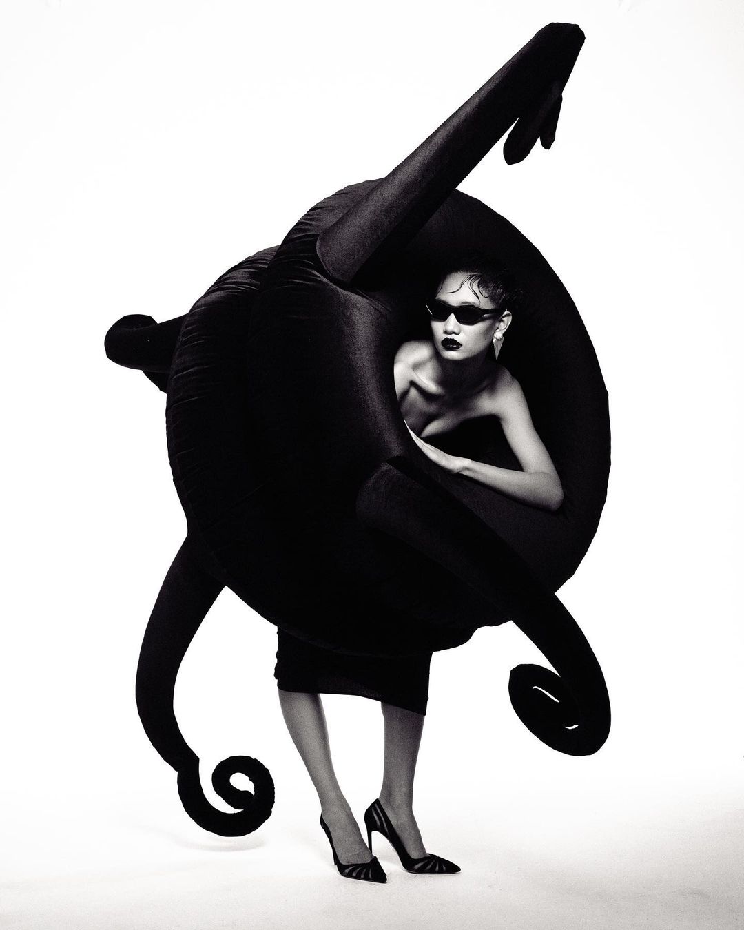 A fashionable woman in sunglasses and heels poses inside a futuristic black plush object against a bright white studio backdrop.