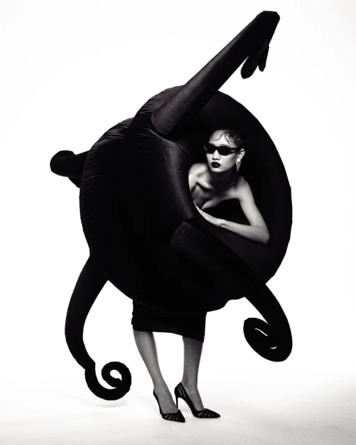 A fashionable woman in sunglasses and heels poses inside a futuristic black plush object against a bright white studio backdrop.