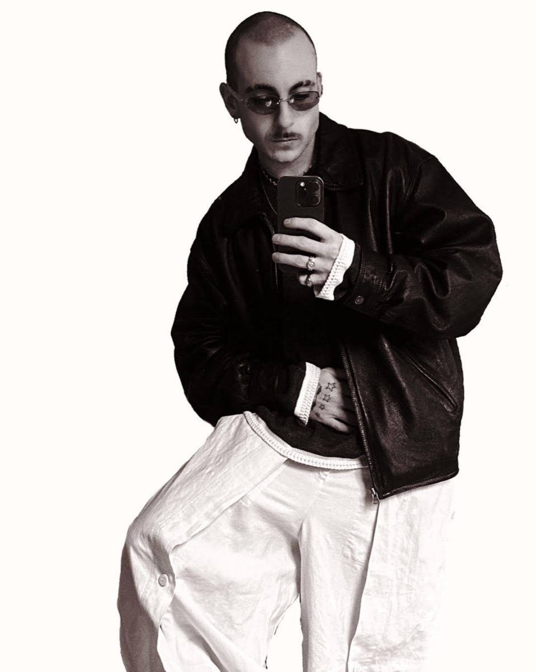An adult male with glasses and mustache takes a selfie. He wears a black jacket, a white shirt, and white pants against a white backdrop.