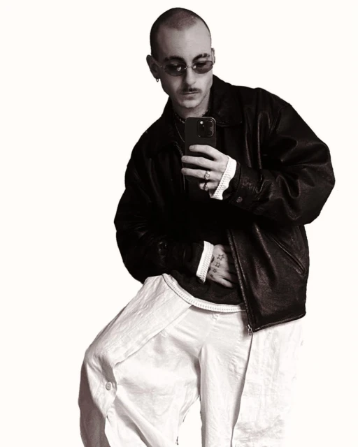 An adult male with glasses and mustache takes a selfie. He wears a black jacket, a white shirt, and white pants against a white backdrop.