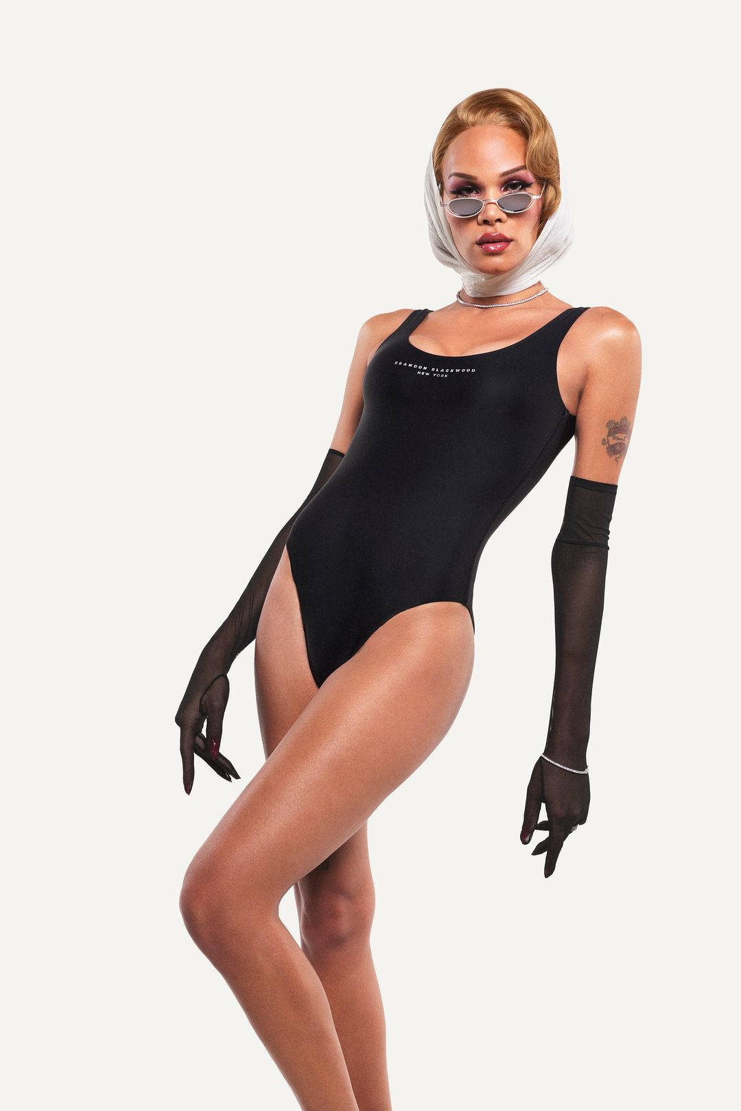 Adult female posing in black swimsuit with sunglasses and sheer gloves in a studio with soft lighting, fashion editorial.