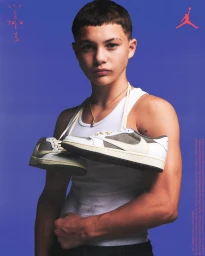 Young man in a white tank top holding sneakers draped over his neck against a solid blue background by photographer Conor Cunningham.