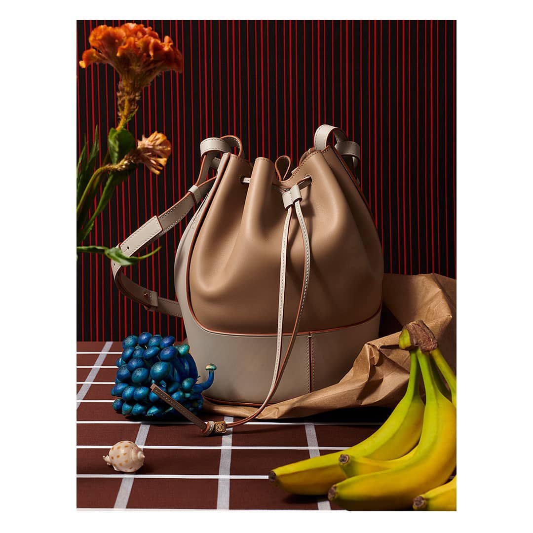 Tan and off-white leather bucket bag on a brown and white checkered surface, styled with bananas, a flower, and a blue sculpture.