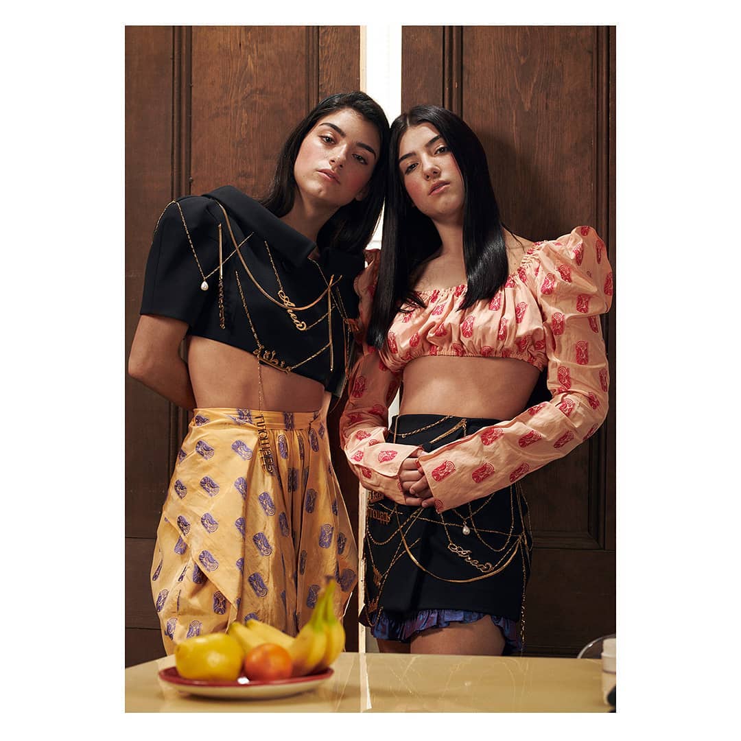 Two young women pose in coordinated outfits indoors; one in black and yellow, the other in pink and black, standing near fruit.