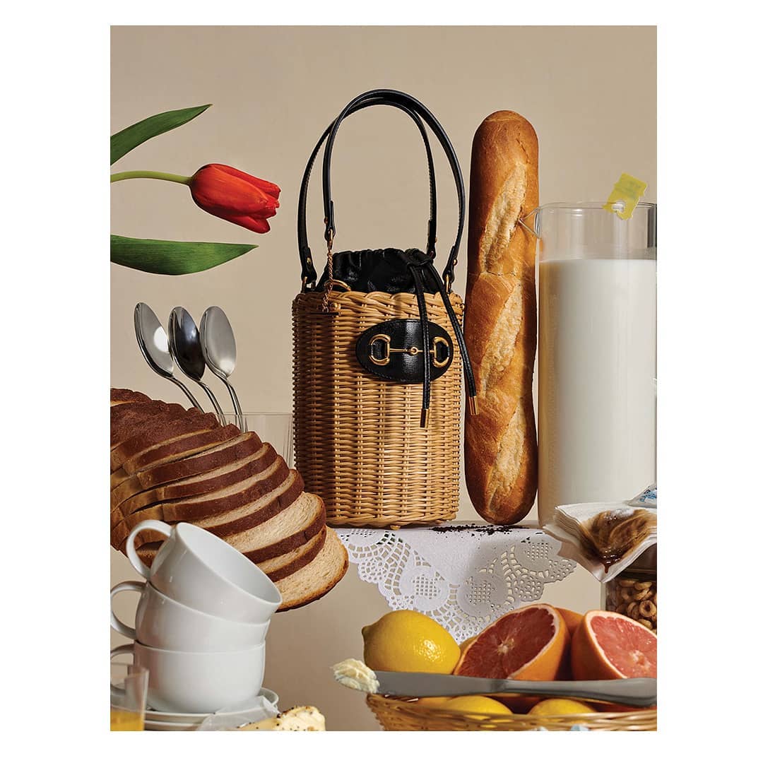 Still life composition features a straw bucket bag, bread loaf, glass of milk, sliced bread, fruit, and silverware against a neutral backdrop.