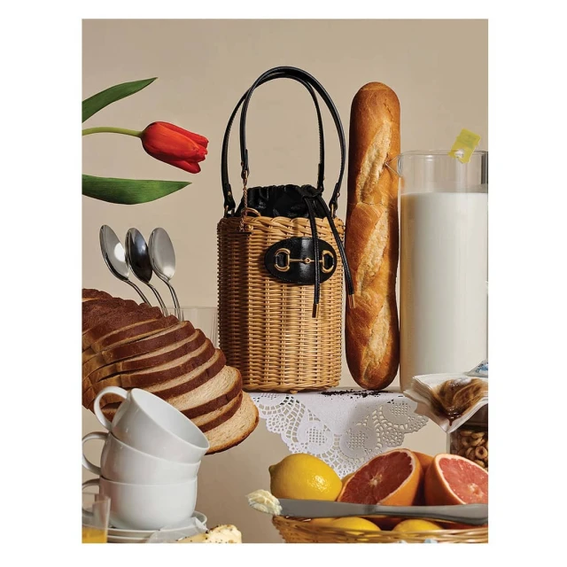 Still life composition features a straw bucket bag, bread loaf, glass of milk, sliced bread, fruit, and silverware against a neutral backdrop.
