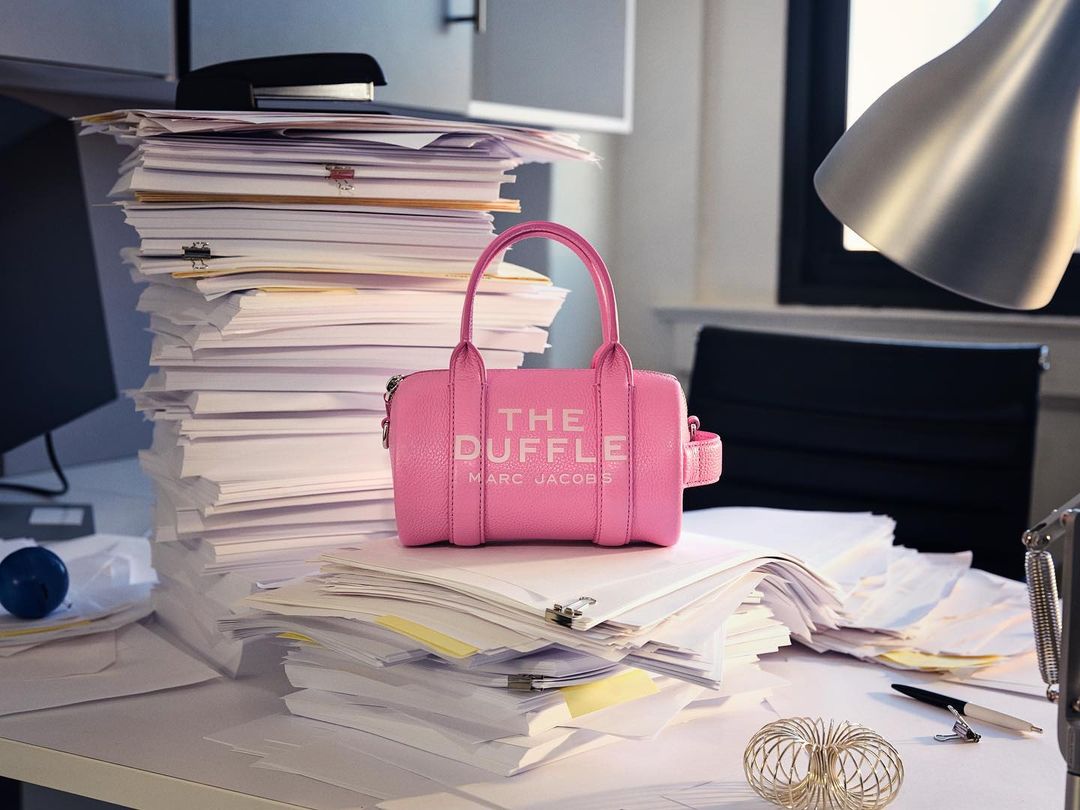 Pink 'The Duffle' Marc Jacobs handbag on a messy white desk covered in stacks of papers and office supplies near a window.