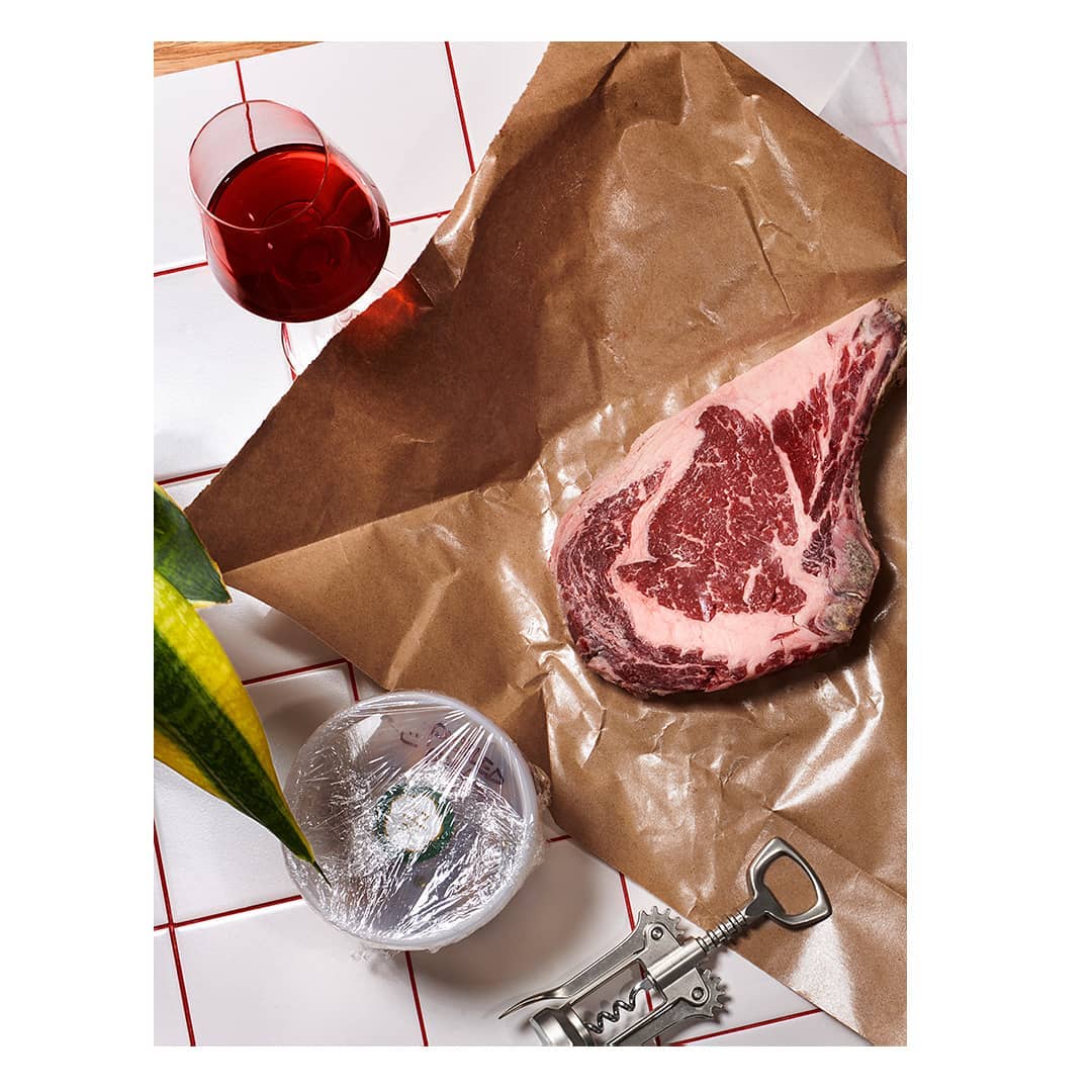 Studio shot of a ribeye steak on brown butcher paper, with wine, a foil-covered bowl, corkscrew, and tiled surface in a modern kitchen setting.