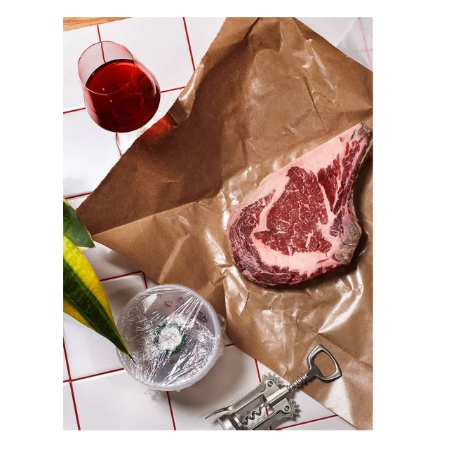 Studio shot of a ribeye steak on brown butcher paper, with wine, a foil-covered bowl, corkscrew, and tiled surface in a modern kitchen setting.