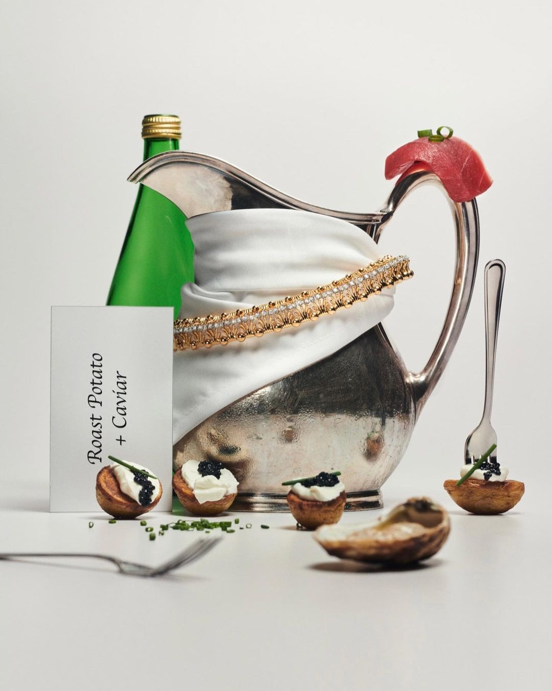 Still life of an elegant silver pitcher with caviar and roast potatoes, complemented by a gold diamond bracelet and green glass bottle.