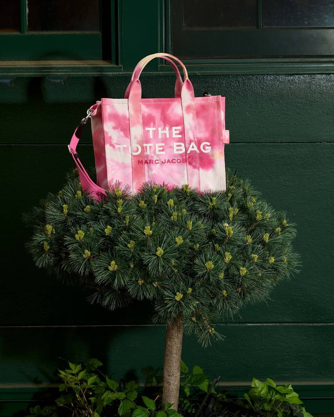 Pink and white tie-dye The Tote Bag by Marc Jacobs is displayed on a trimmed evergreen topiary against a dark green door.