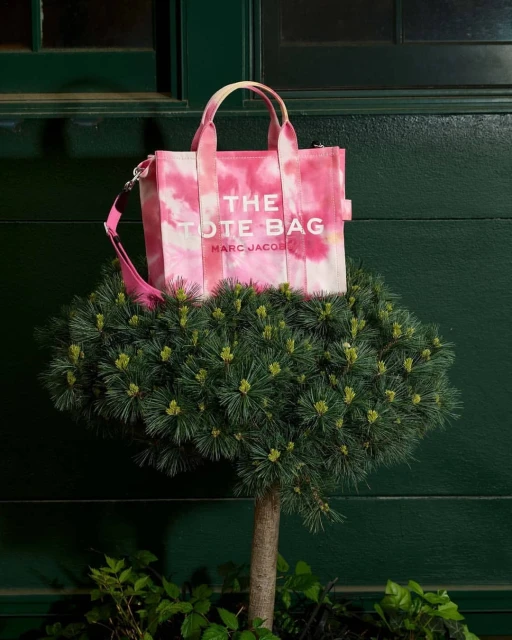 Pink and white tie-dye The Tote Bag by Marc Jacobs is displayed on a trimmed evergreen topiary against a dark green door.