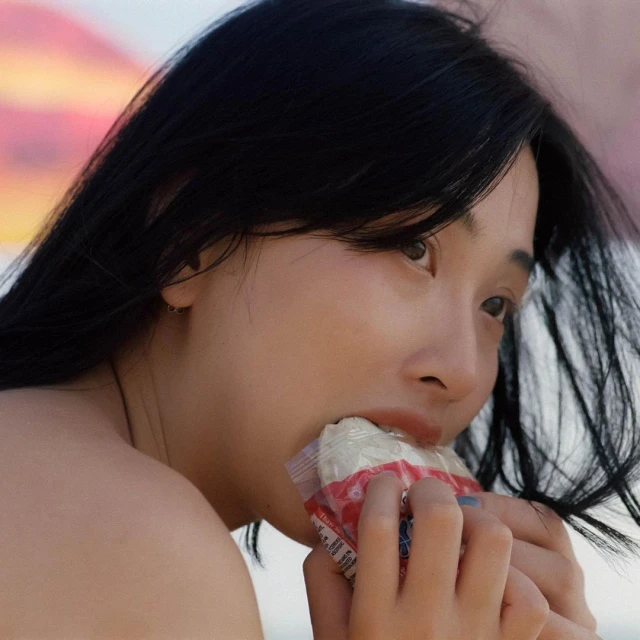 Close up of a young Asian woman with dark hair eating a snack from a plastic wrapper outside on a sunny day in natural light.