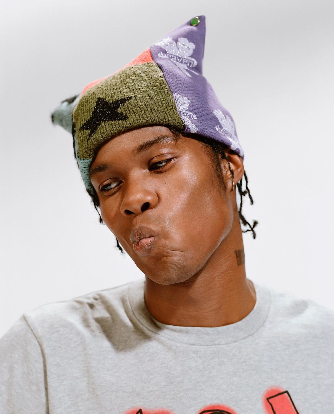 Portrait of a young adult with dark skin wearing a colorful knit hat against a plain white backdrop, looking off-camera.