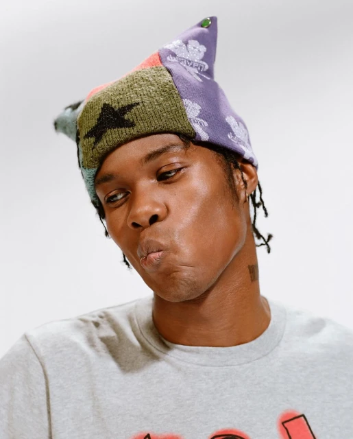 Portrait of a young adult with dark skin wearing a colorful knit hat against a plain white backdrop, looking off-camera.