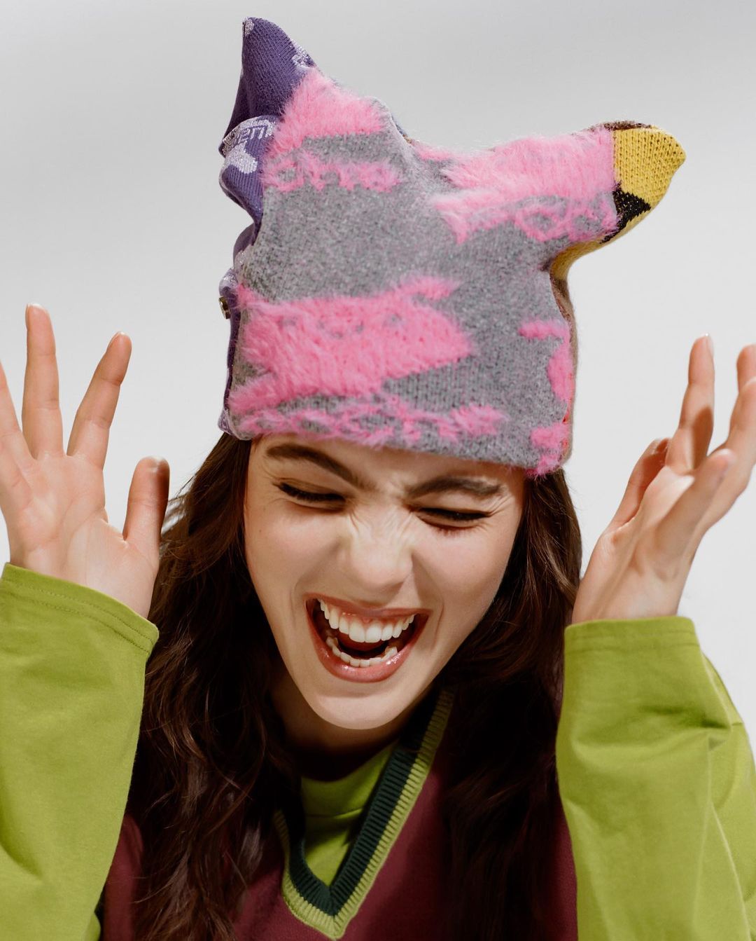 Smiling woman wearing a colorful knitted hat, green long-sleeved shirt, and burgundy sweater vest, set against a plain white background.