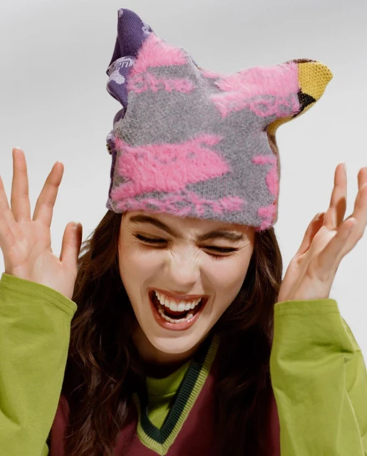 Smiling woman wearing a colorful knitted hat, green long-sleeved shirt, and burgundy sweater vest, set against a plain white background.