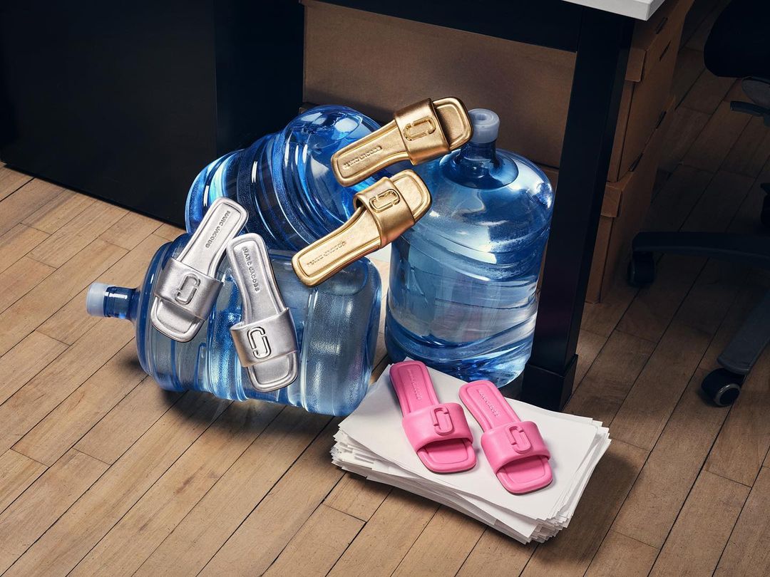 Eye-level shot of gold, silver, and pink slide sandals placed on water bottles, a stack of paper, and a wooden floor.