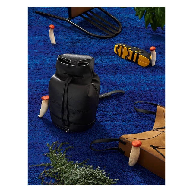 Leather backpack on a blue shag rug, surrounded by leather accessories and small mushroom-shaped objects with green vegetation in view.