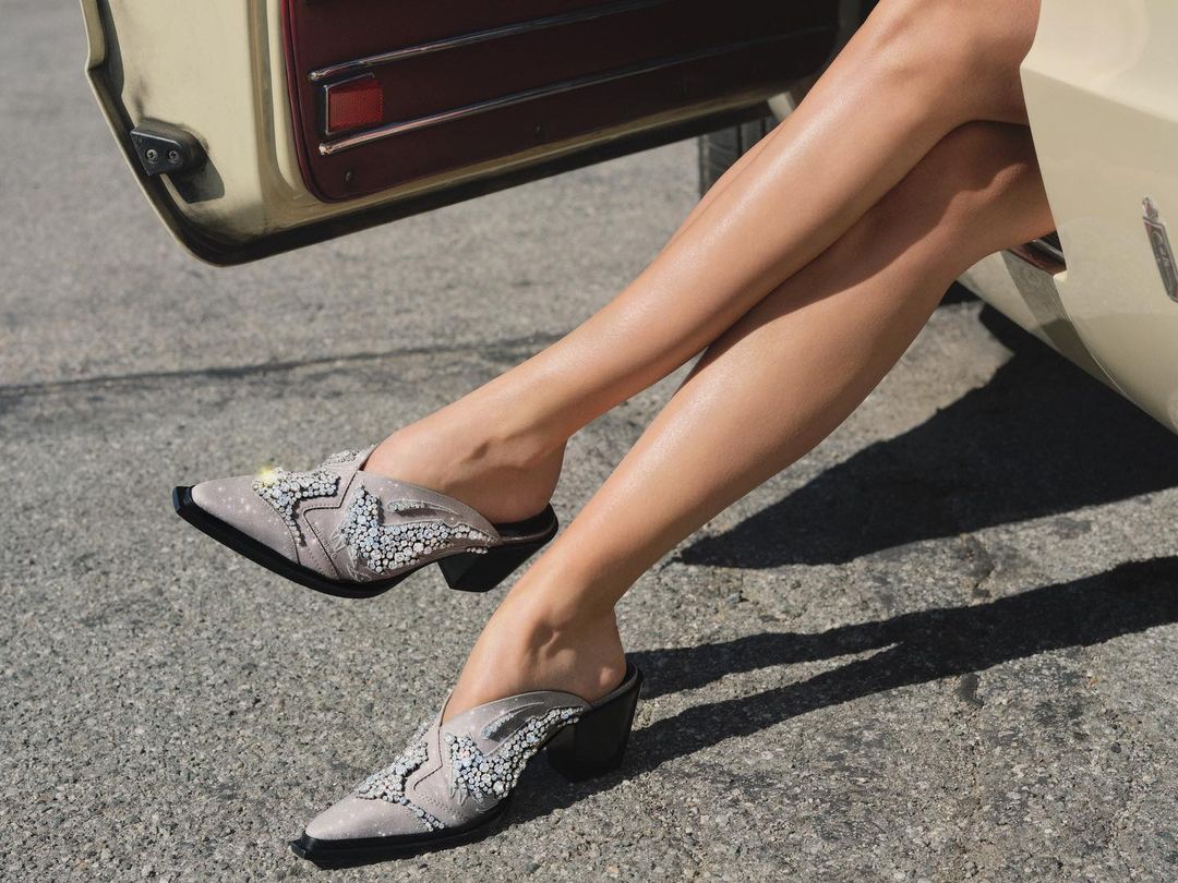 Elegant mule shoes with black block heels and decorative beading are featured on a woman sitting in a vintage car.