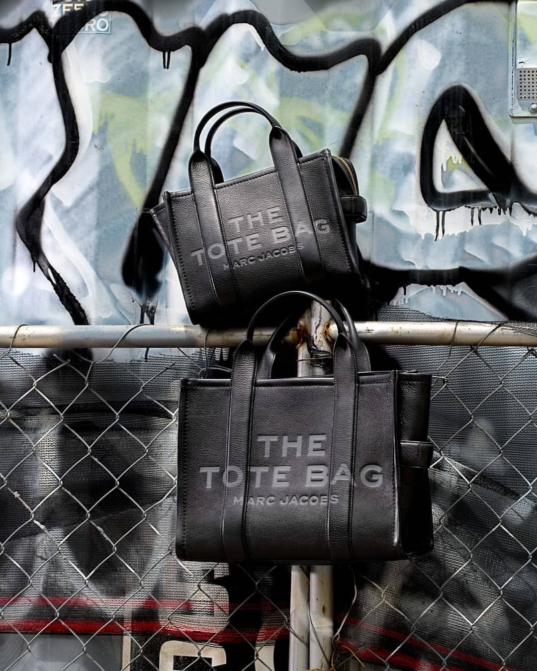 Two black leather Marc Jacobs 'The Tote Bags' are displayed on a metal railing with a graffiti-covered wall in the background.