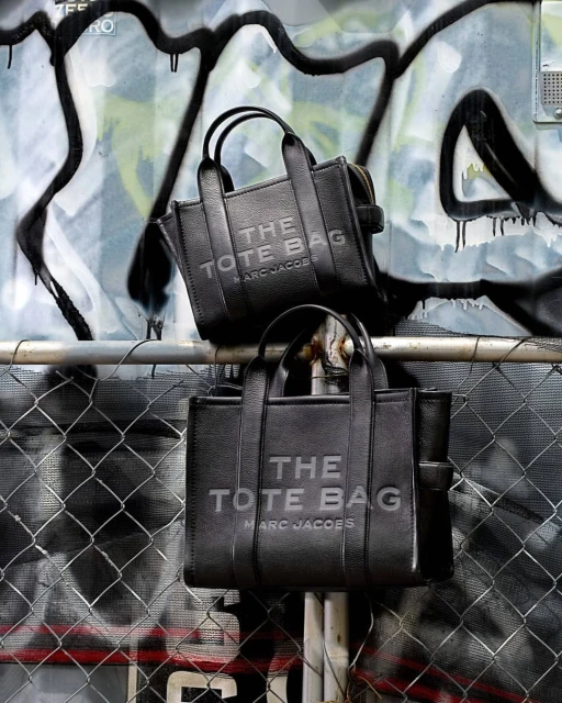 Two black leather Marc Jacobs 'The Tote Bags' are displayed on a metal railing with a graffiti-covered wall in the background.