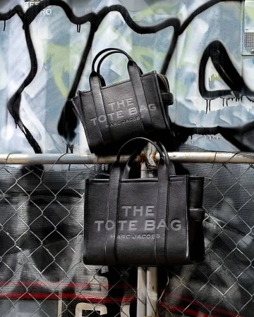 Two black leather Marc Jacobs 'The Tote Bags' are displayed on a metal railing with a graffiti-covered wall in the background.