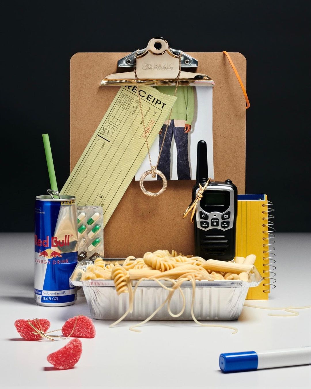 Still life of pasta in aluminum pan, can of Red Bull, receipt, jewelry, and pills on white surface with dark background.