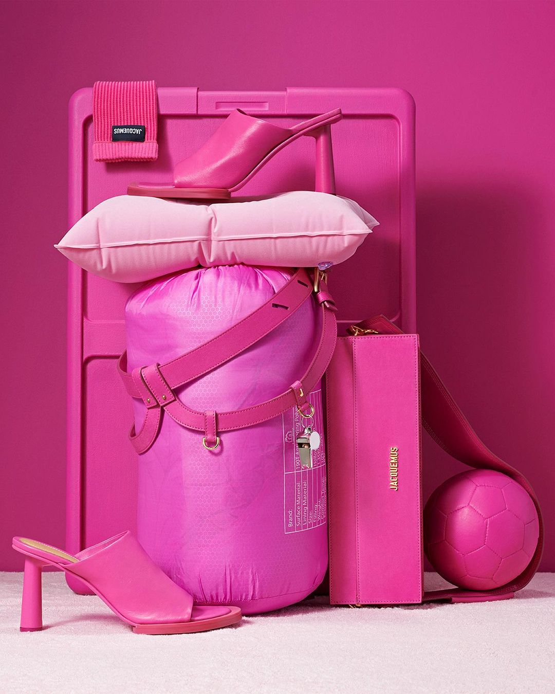 Still life arrangement of pink accessories, including high heel shoes, a tote bag, a beach towel, and a soccer ball against a pink backdrop.