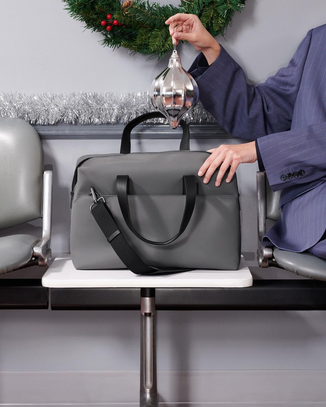 Gray duffel bag on a white table with a person in a suit holding an ornament in an indoor waiting area with festive Christmas decorations.