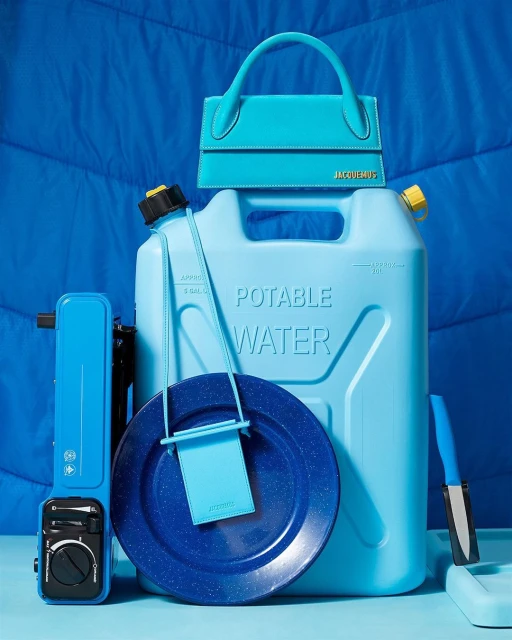 Still life studio shot of turquoise Jacquemus handbags atop a matching water jug, plate, camping stove, and blue quilted backdrop.