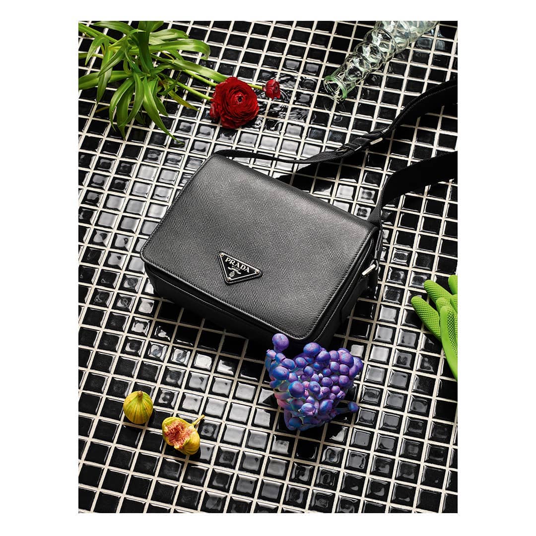 Prada black leather messenger bag with figs, a red flower, and a glass on a black and white tiled surface. Fashion product shot.