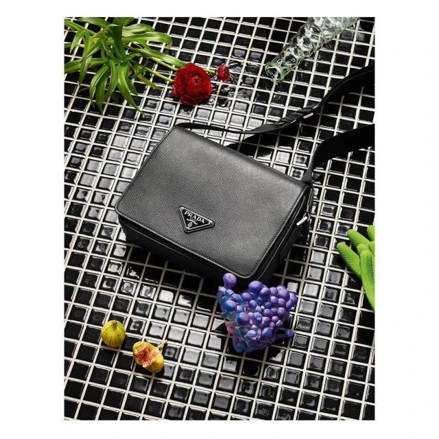 Prada black leather messenger bag with figs, a red flower, and a glass on a black and white tiled surface. Fashion product shot.