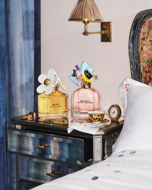 Two Marc Jacobs perfume bottles displayed on a reflective nightstand next to a bed, illuminated by a wall sconce in an indoor setting.