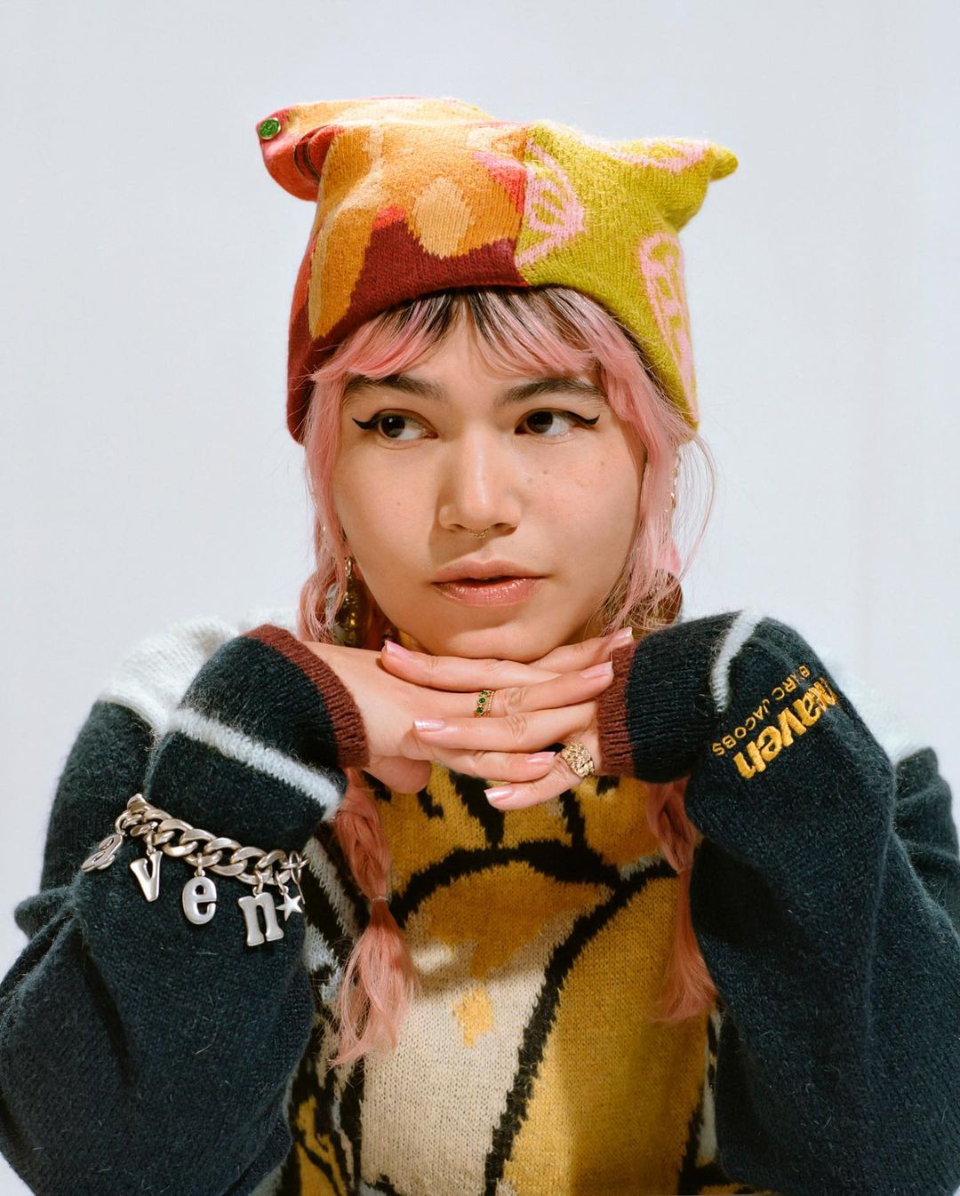 Portrait of a pink-haired young adult woman wearing a graphic sweater and patterned hat against a white studio backdrop, fashion shoot.