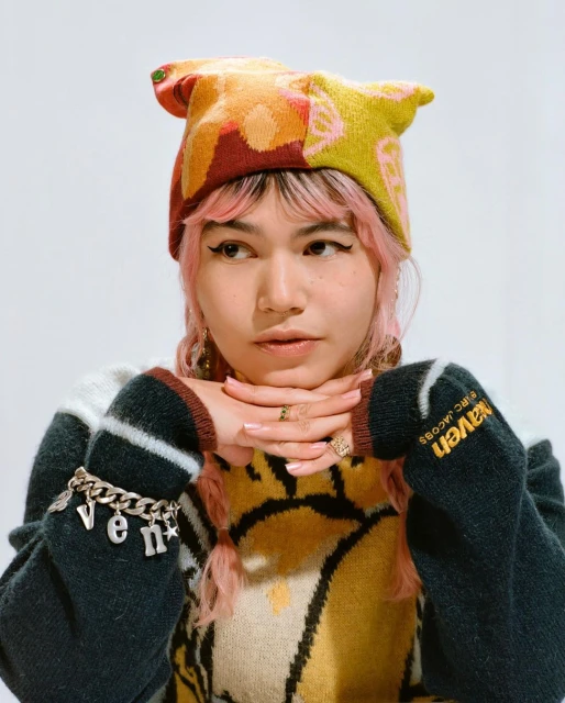 Portrait of a pink-haired young adult woman wearing a graphic sweater and patterned hat against a white studio backdrop, fashion shoot.
