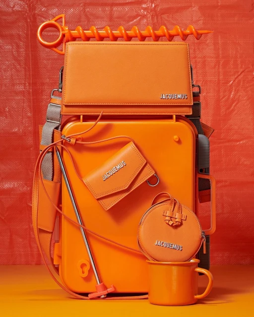 Still life with orange Jacquemus bags, a matching mug, and a large plastic toy auger against a textured orange backdrop. Fashion-forward studio shot.