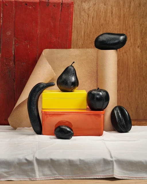 Still life of glossy black fruit and colorful boxes against a backdrop of brown paper and a red wooden panel on a white tablecloth.